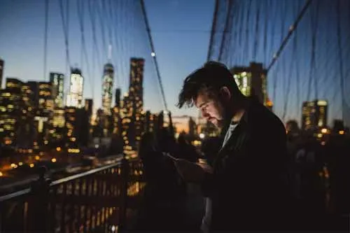 Người đàn ông nhìn vào điện thoại của mình khi đứng trên Cầu Brooklyn lúc hoàng hôn với nền đường chân trời của thành phố