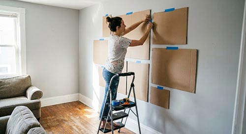 Ảnh chụp thực tế một người đang dùng băng dính giấy hoặc bìa carton dán lên tường để ướm thử kích thước tranh.